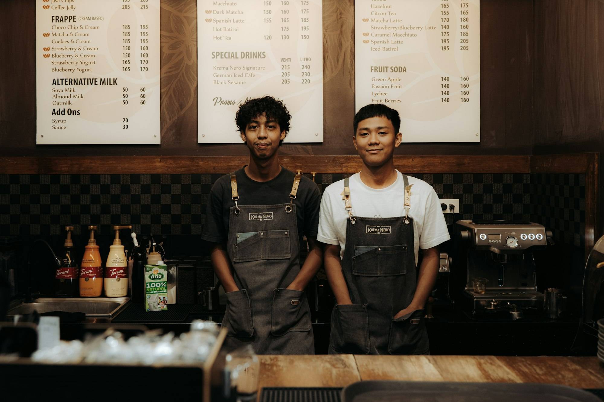 Two cafe owners in aprons smiling behind the counter of their coffee shop